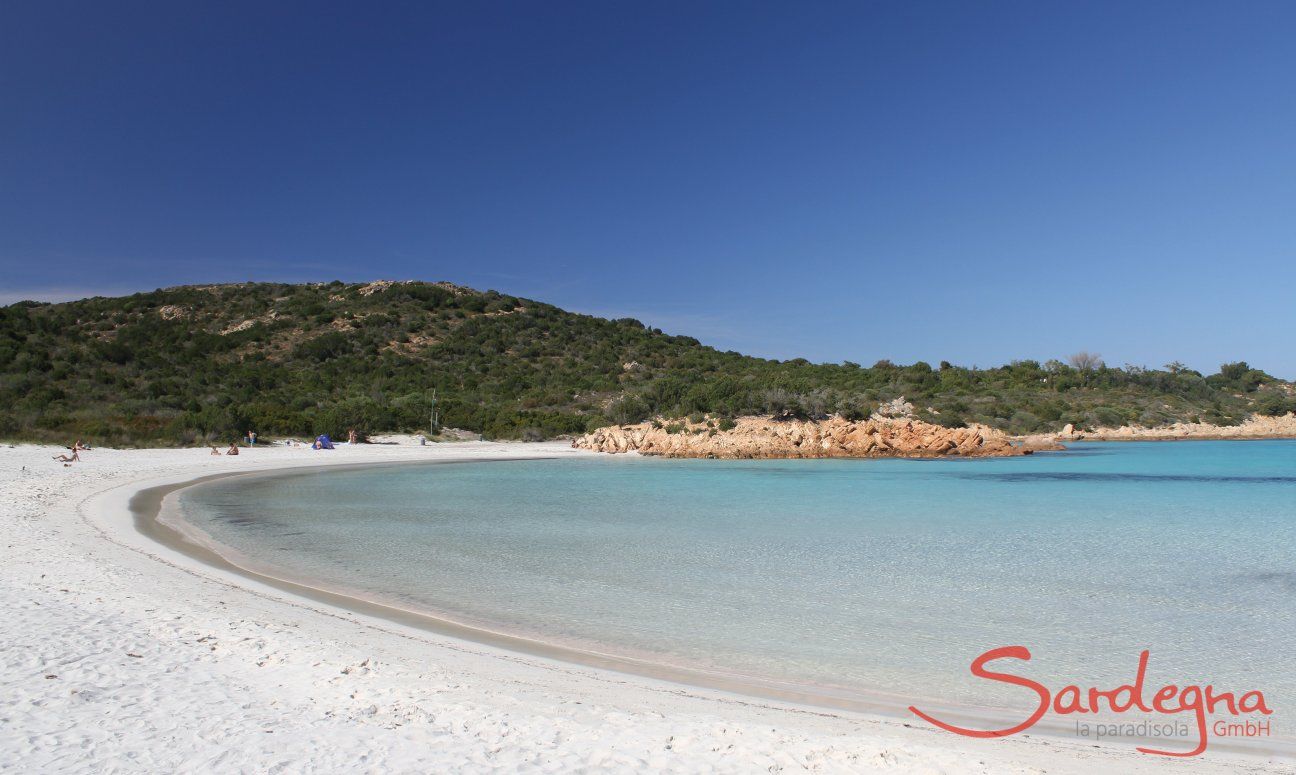 Spiaggia Romazzino fatto di sabbia bianca a forma di mezza luna, alla fine rocce giallie e una collina coperta di macchia verde